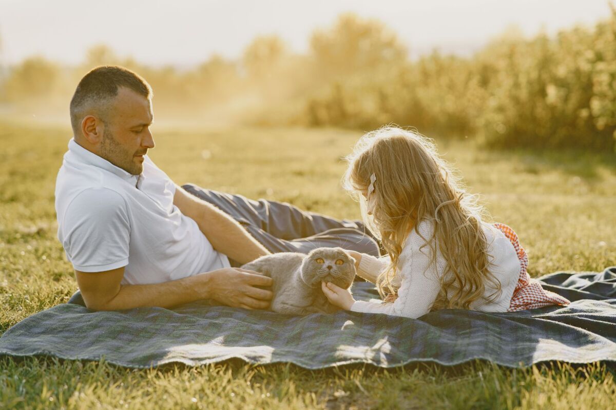 A father and daughter relaxing outdoors with a Scottish Fold cat during a sunny day.