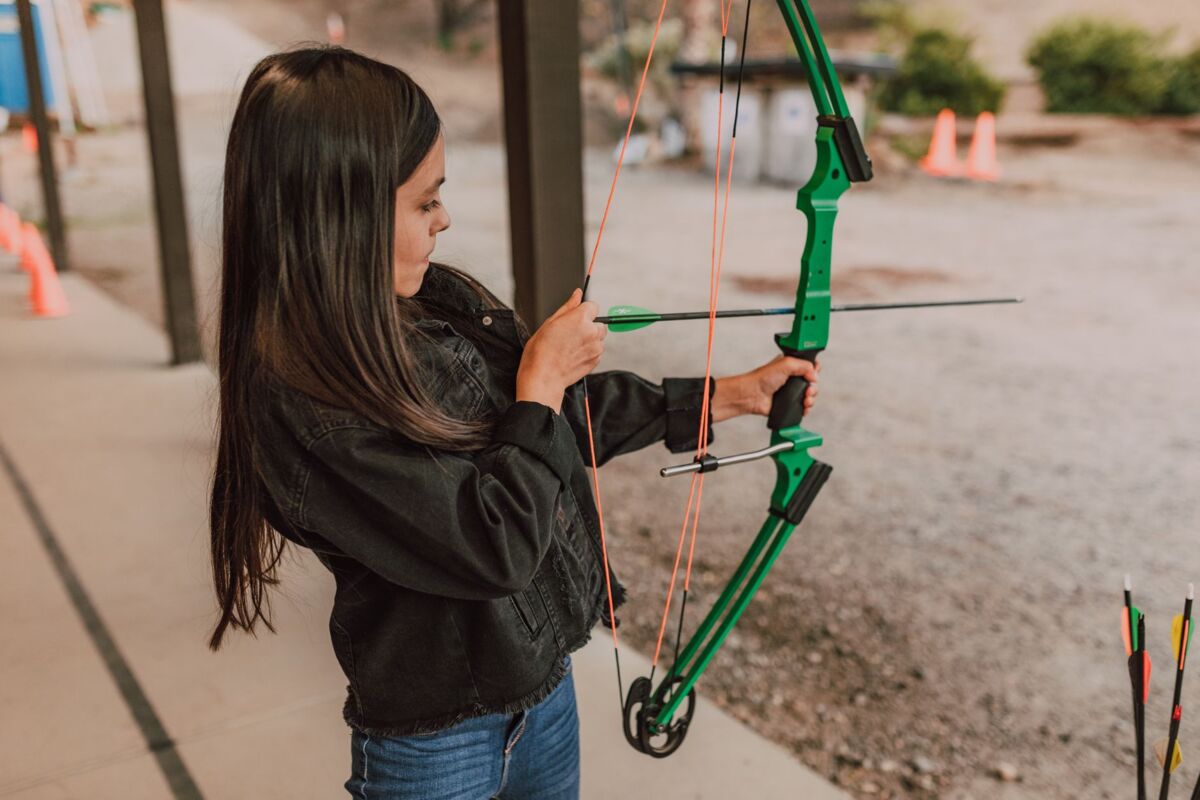 Girl practicing archery, aiming with a green bow at an outdoor range.