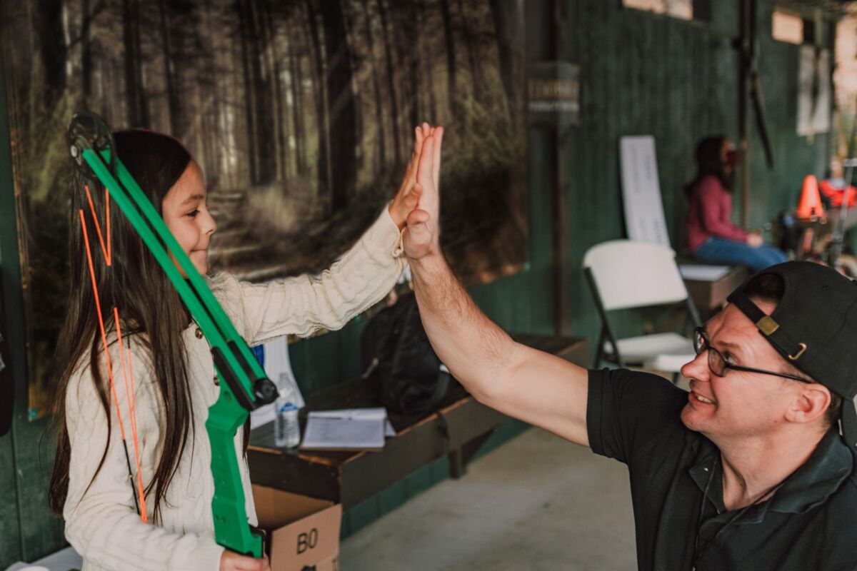A young girl and her father share a high five during an archery session at an indoor range.