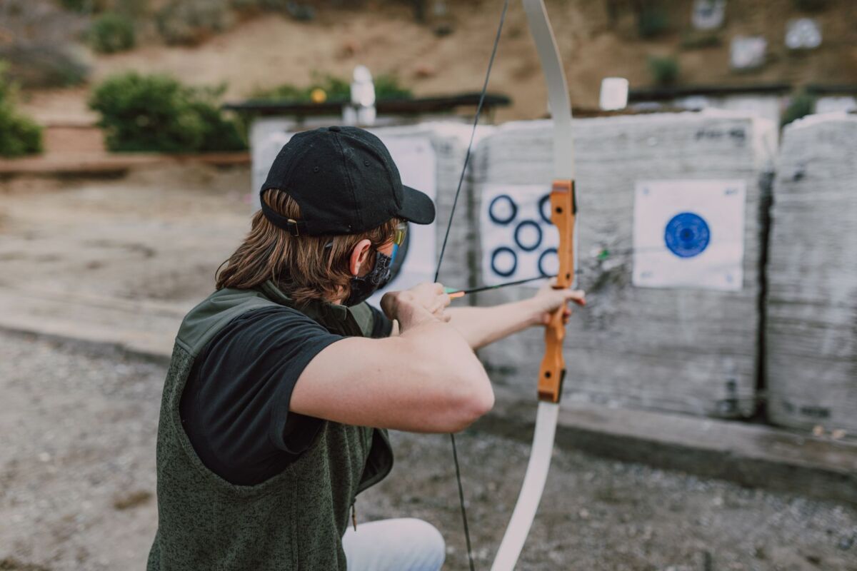 Young adult practicing archery with a recurve bow at an outdoor range on a sunny day.