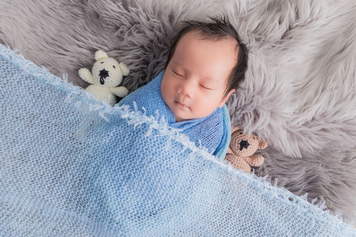 Asian baby sleeping soundly, wrapped in a blue blanket with stuffed toys.