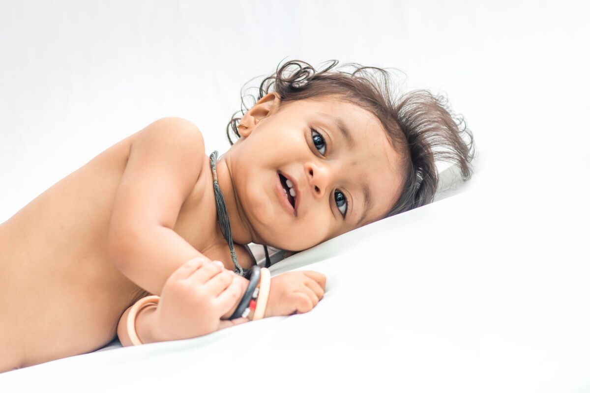 Cute South Asian baby lying on white bed with bright eyes and playful expression.