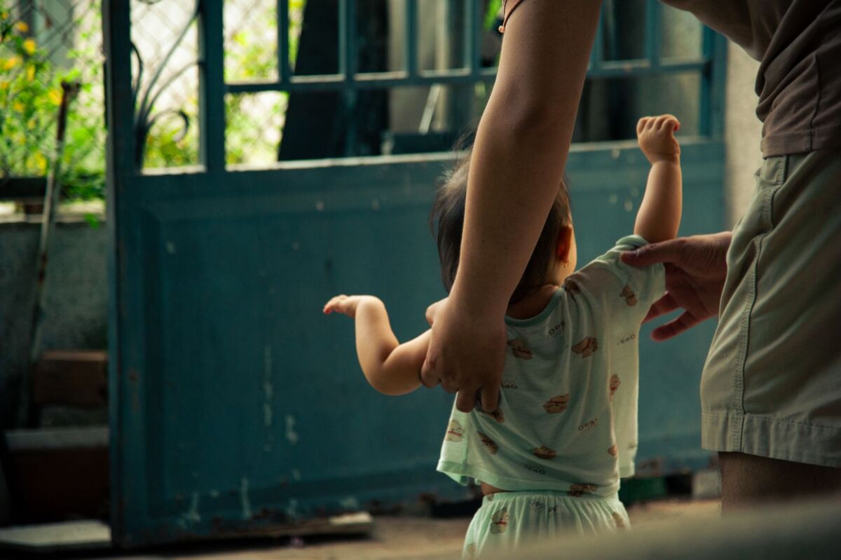 A tender moment of a toddler learning to walk indoors with adult support.
