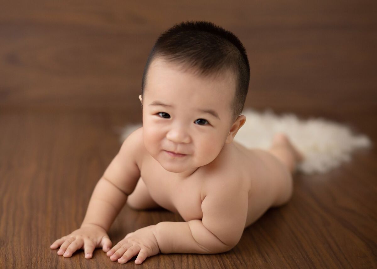Delighted Asian little naked child lying on wooden floor in studio and looking at camera
