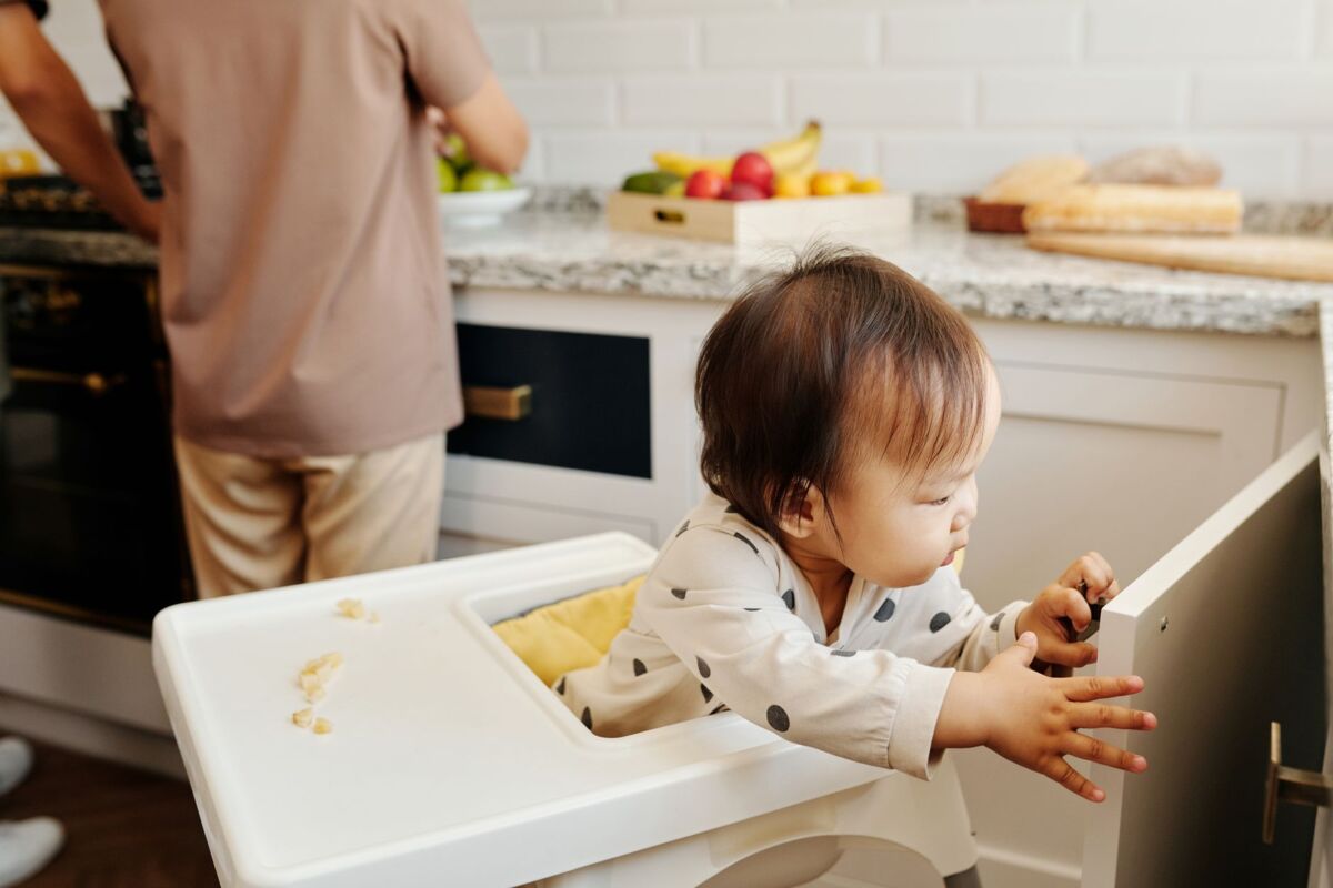 Adorable baby in high chair exploring a kitchen cabinet while parent cooks nearby.