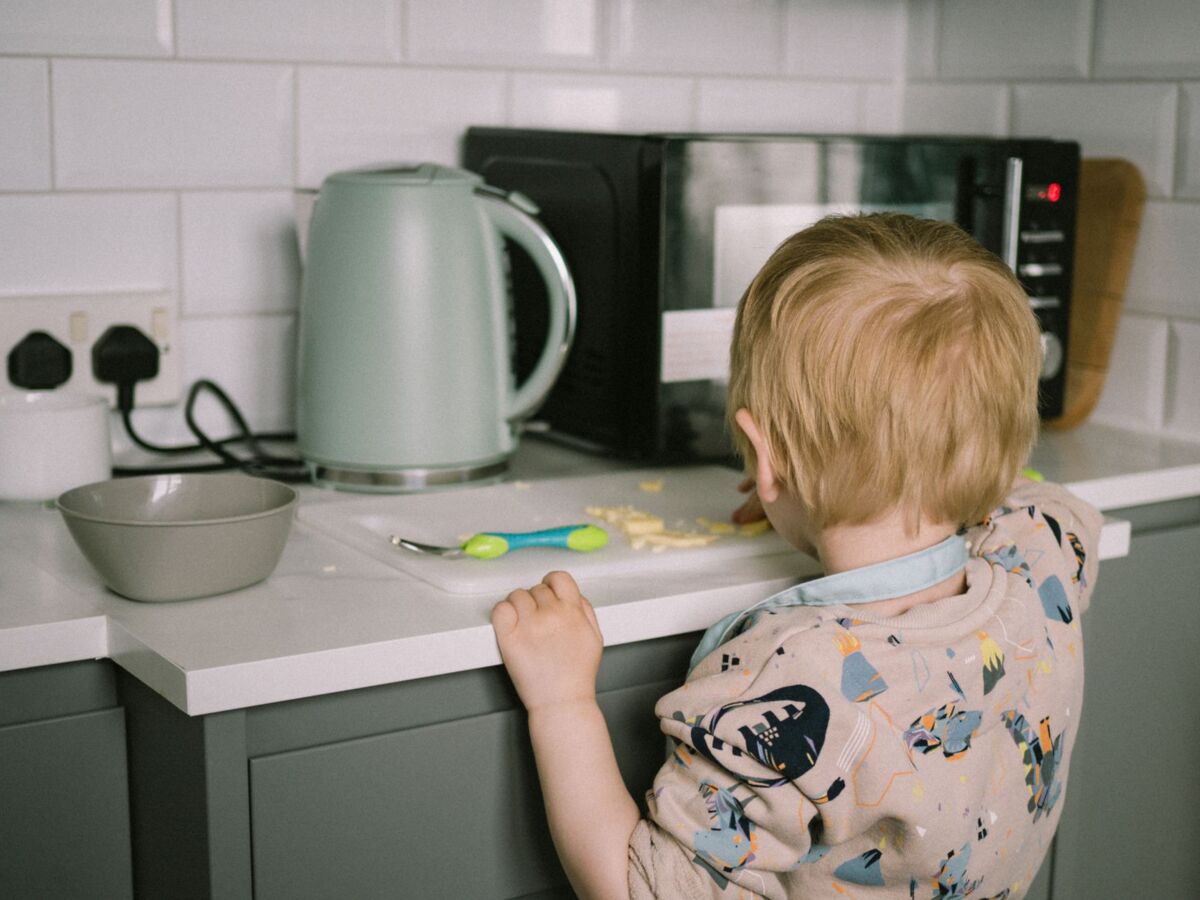 A toddler reaching for food on the kitchen counter next to a kettle and microwave.