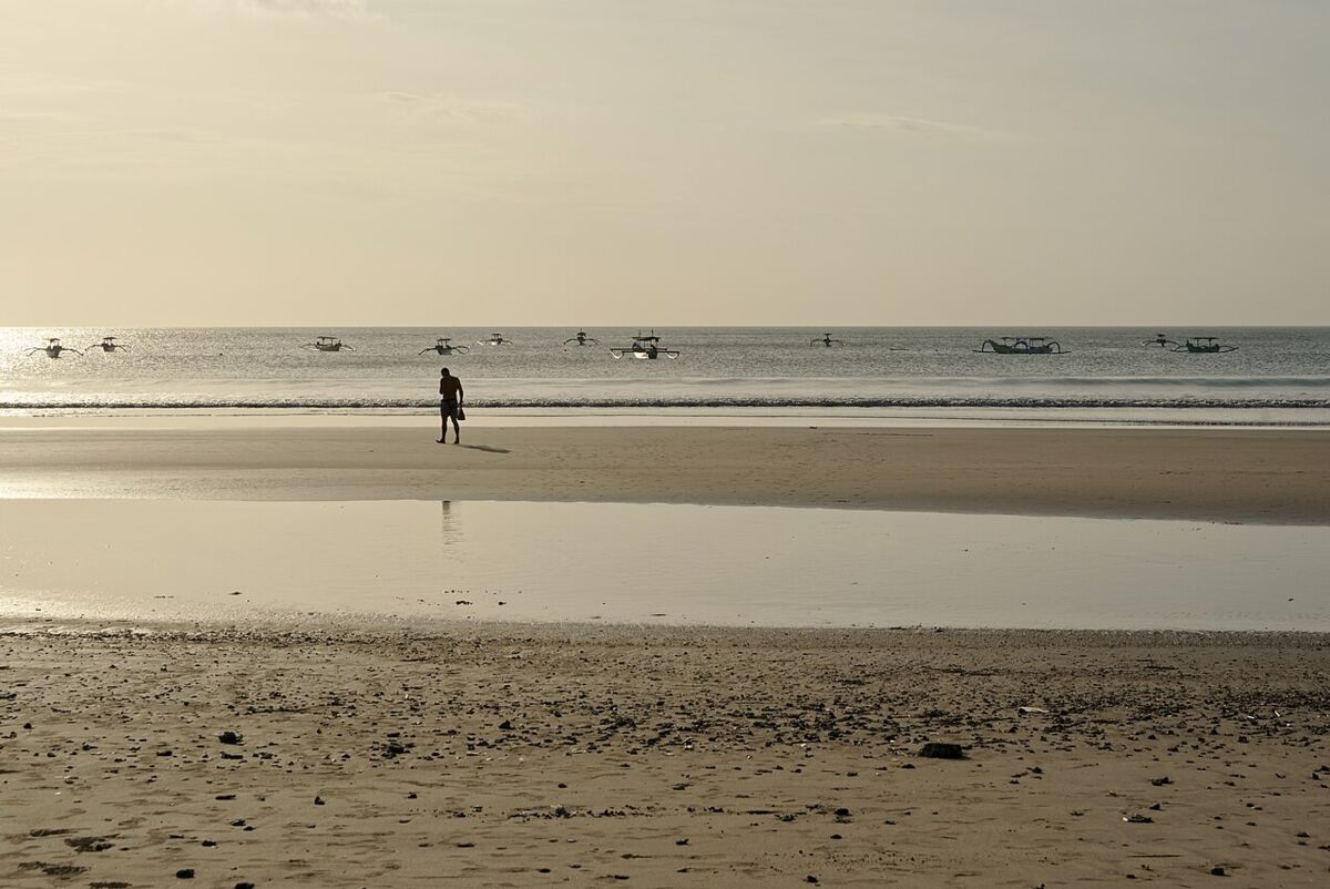 Person walking on a calm reflective beach at low tide with traditional jukung boats offshore, Kuta, Bali