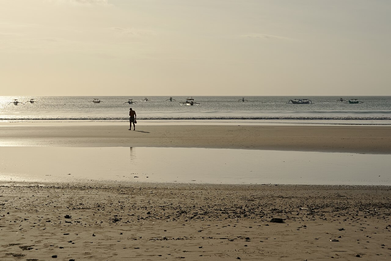 Person walking on a calm reflective beach at low tide with traditional jukung boats offshore, Kuta, Bali