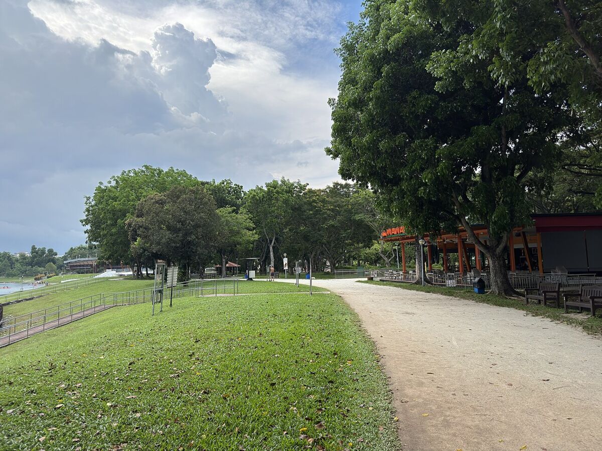 Waterfront walking path at Bedok Reservoir Park beside an open-air eatery, with lawns, trees and the reservoir under a cloudy sky