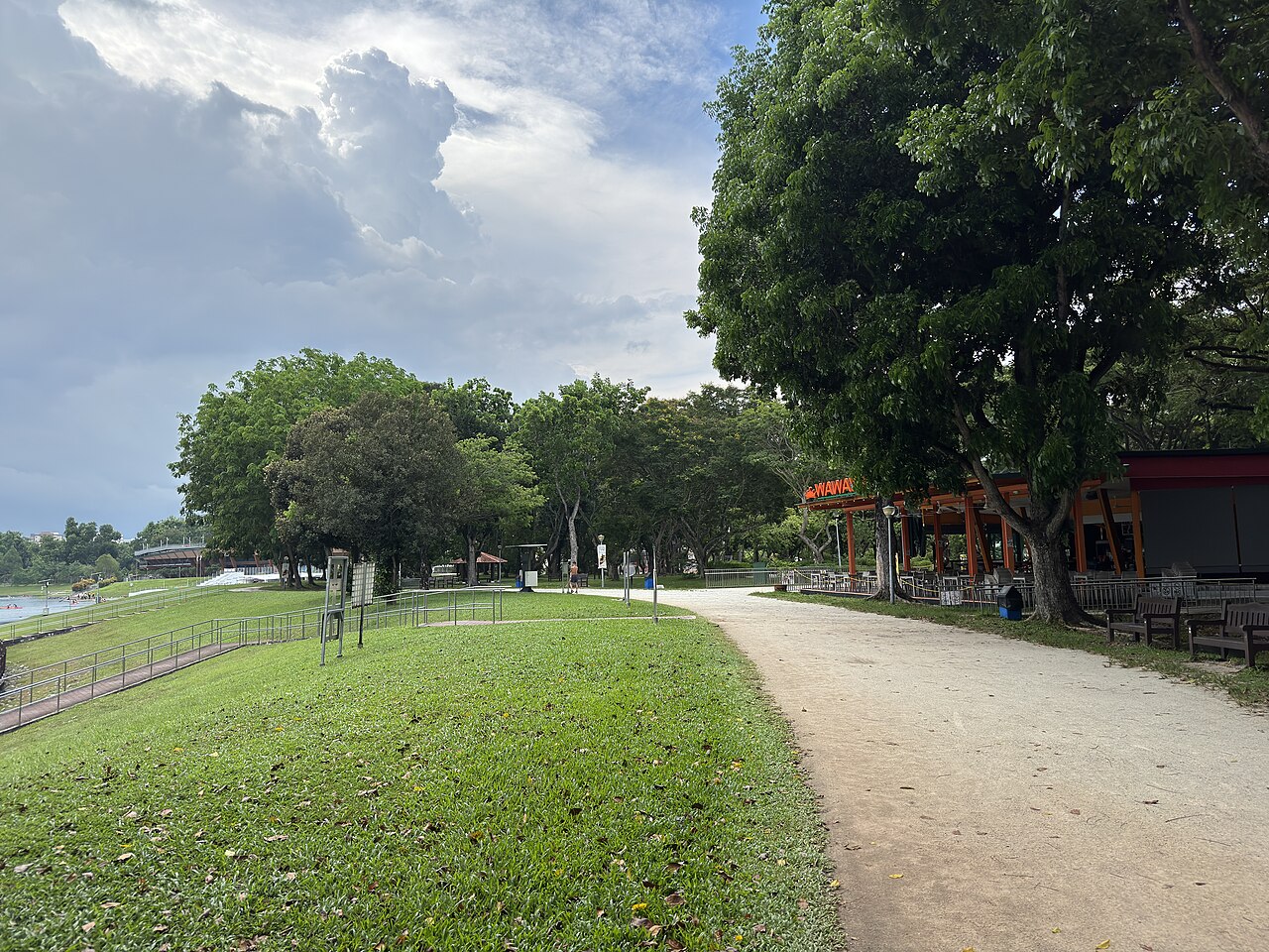 Waterfront walking path at Bedok Reservoir Park beside an open-air eatery, with lawns, trees and the reservoir under a cloudy sky
