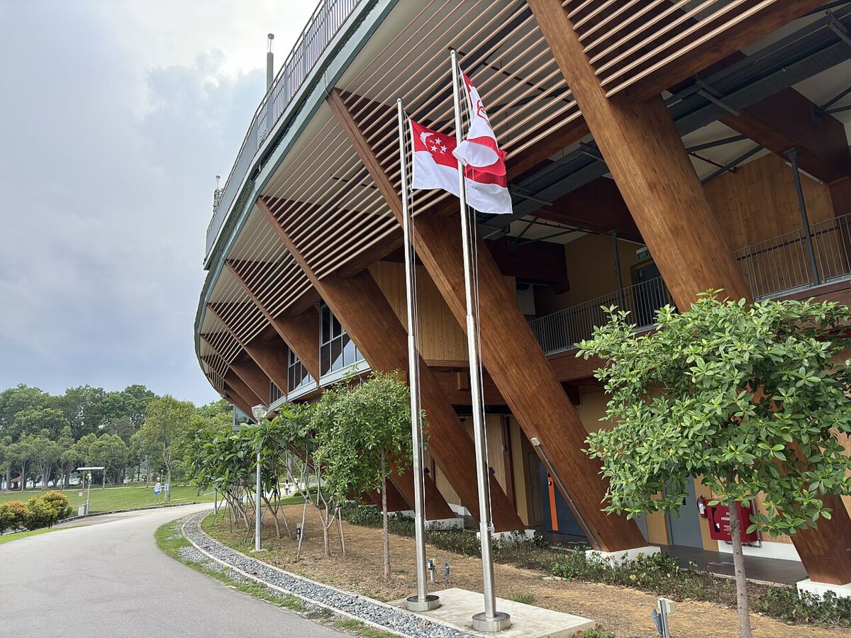 Modern curved timber building with Singapore flags at Bedok Reservoir Park, beside a landscaped path