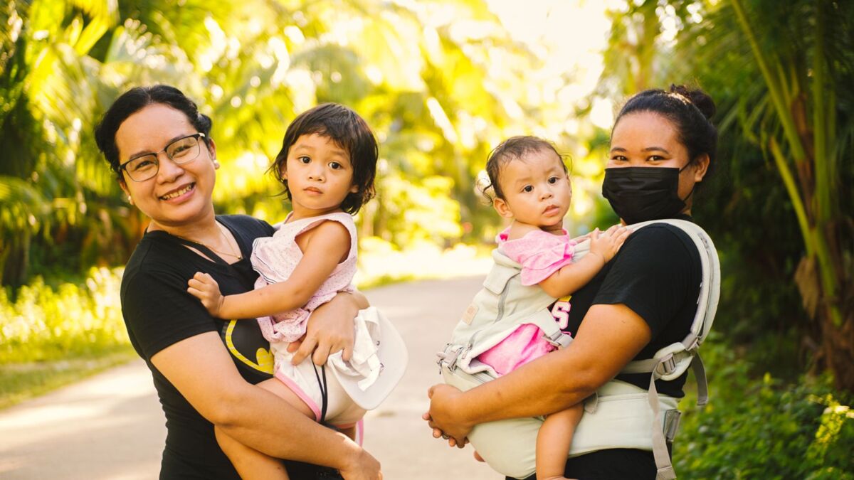 Two mothers happily carry their children outside in a sunny, lush green environment.