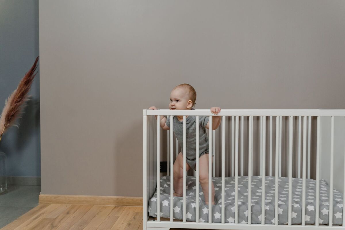 A cute baby in a grey onesie stands in a white crib indoors.