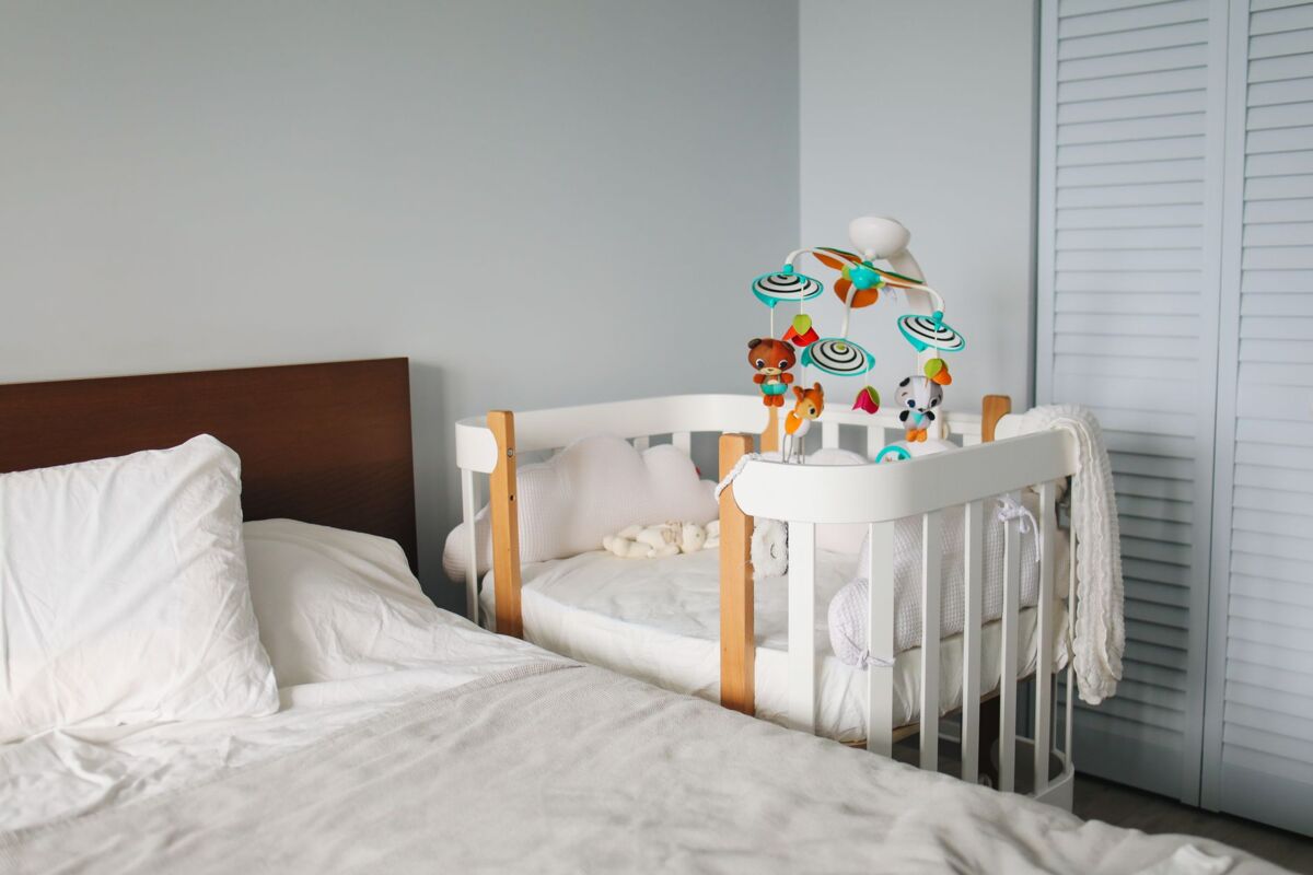 Cozy bedroom with white cute wooden crib locating in corner near empty bed with white sheet and pillows and built in wardrobe on daytime