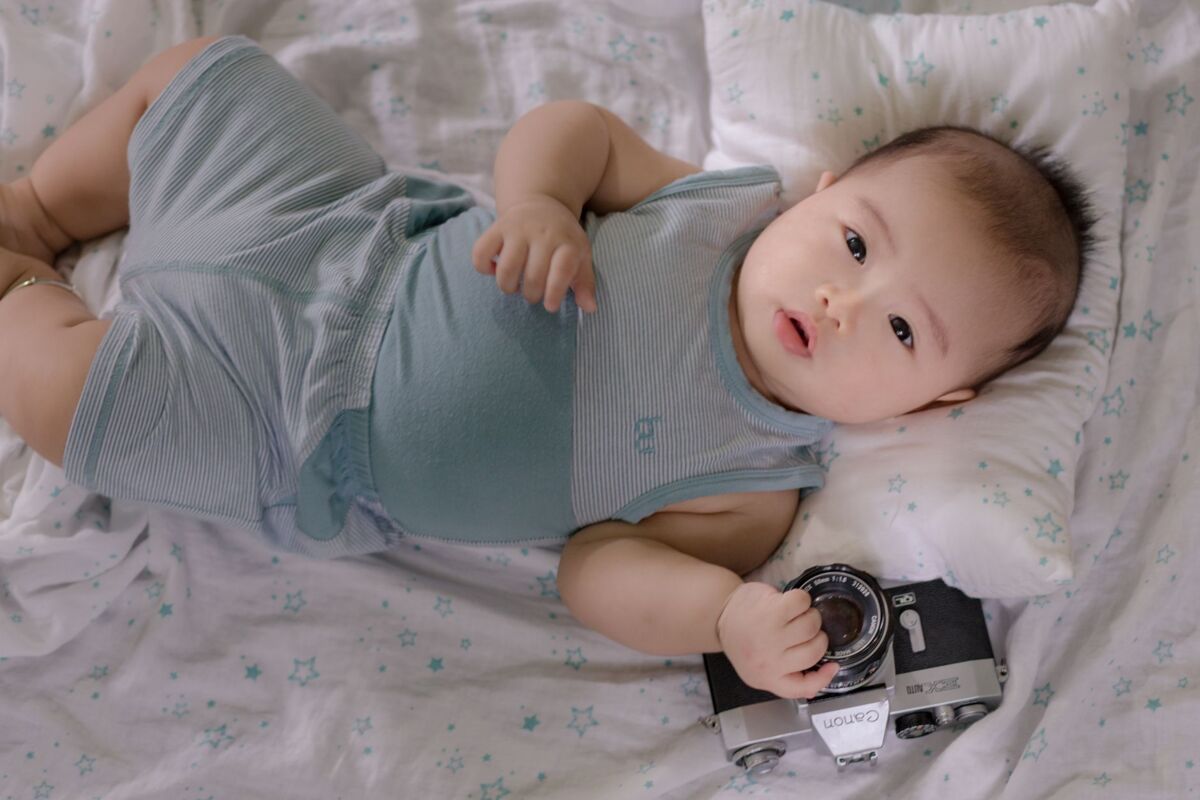 Top view of cute infant Asian child holding photo camera while lying on soft bed in nursery