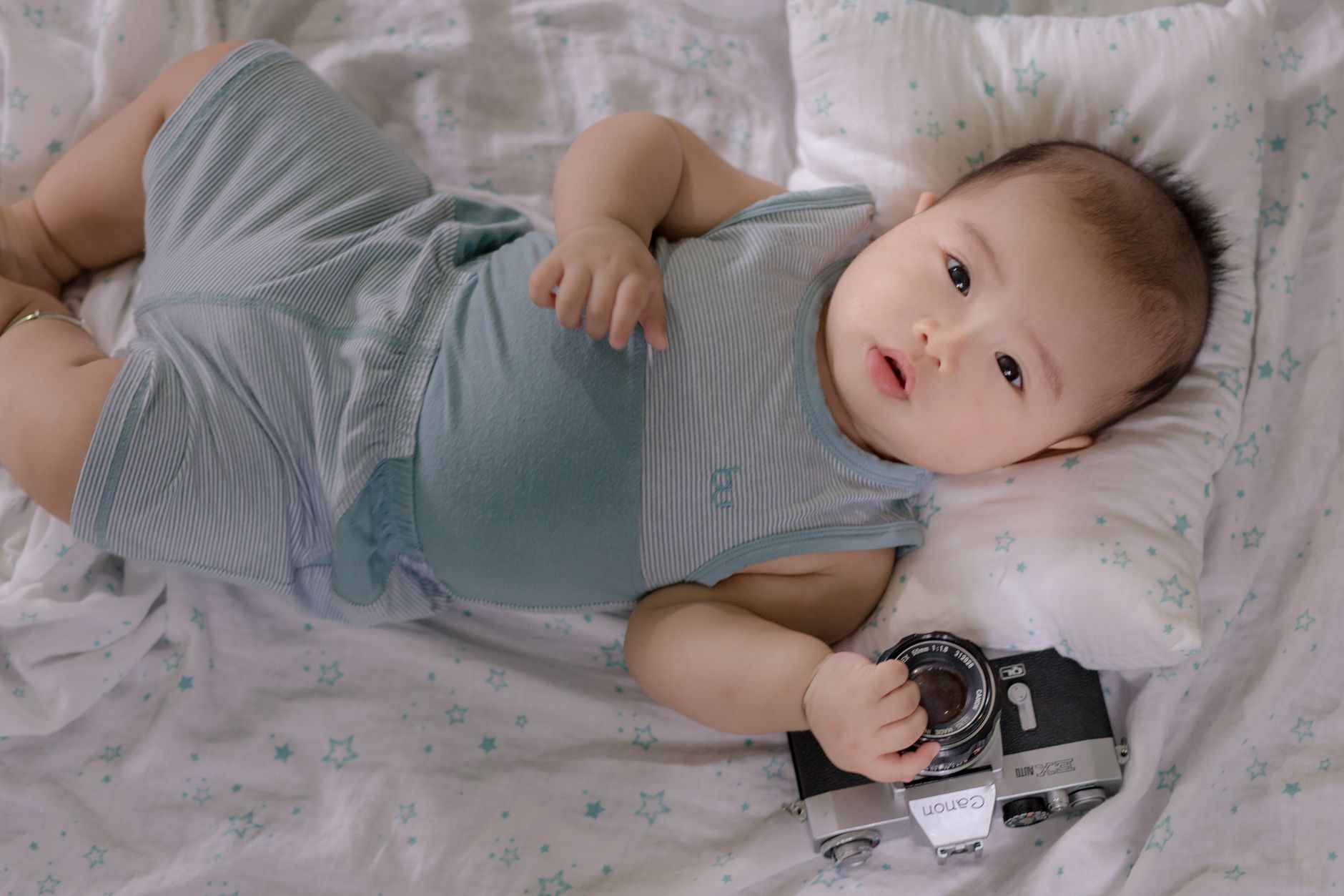 Top view of cute infant Asian child holding photo camera while lying on soft bed in nursery