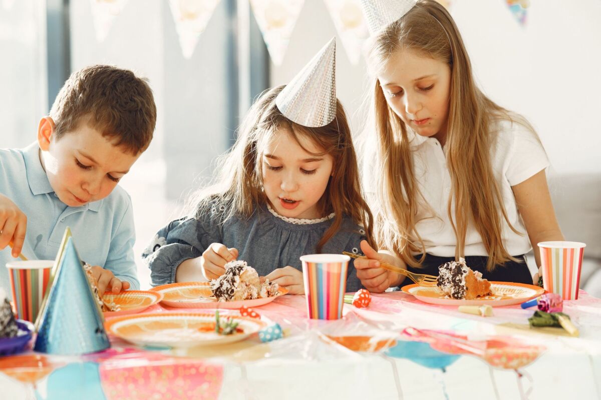 Three children enjoying cake at a colorful birthday party with hats and decorations.