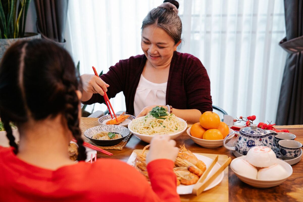 Cheerful ethnic grandma with cooked prawn against anonymous female teen at table with assorted tasty dishes at home