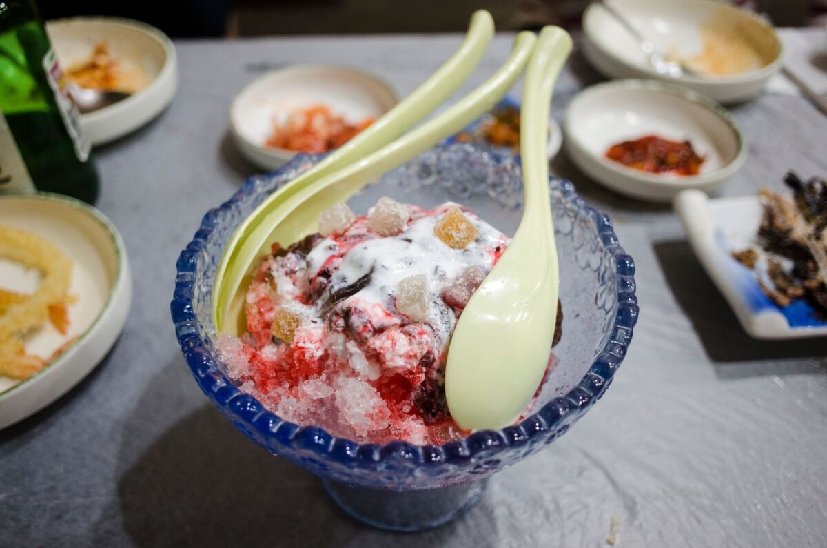 Close-up of a colorful Korean bingsu dessert with spoons in an indoor dining setting.