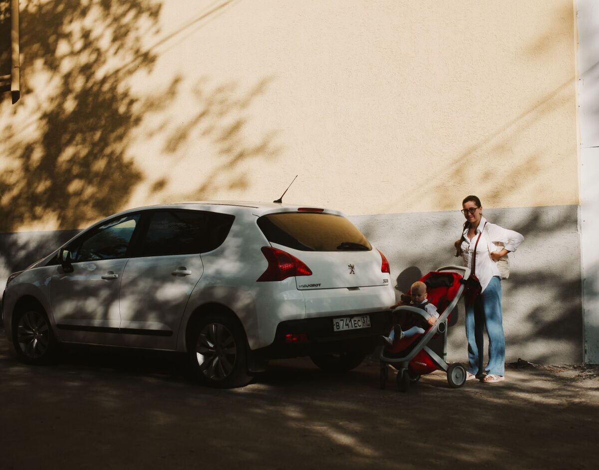 Mother with stroller and child beside a white car on a sunlit street.