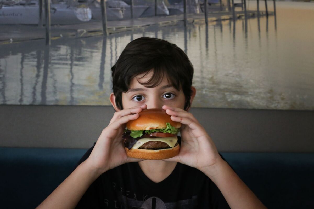 A child holds a delicious cheeseburger, ready to eat, seated indoors with a serene waterfront scene in the background.