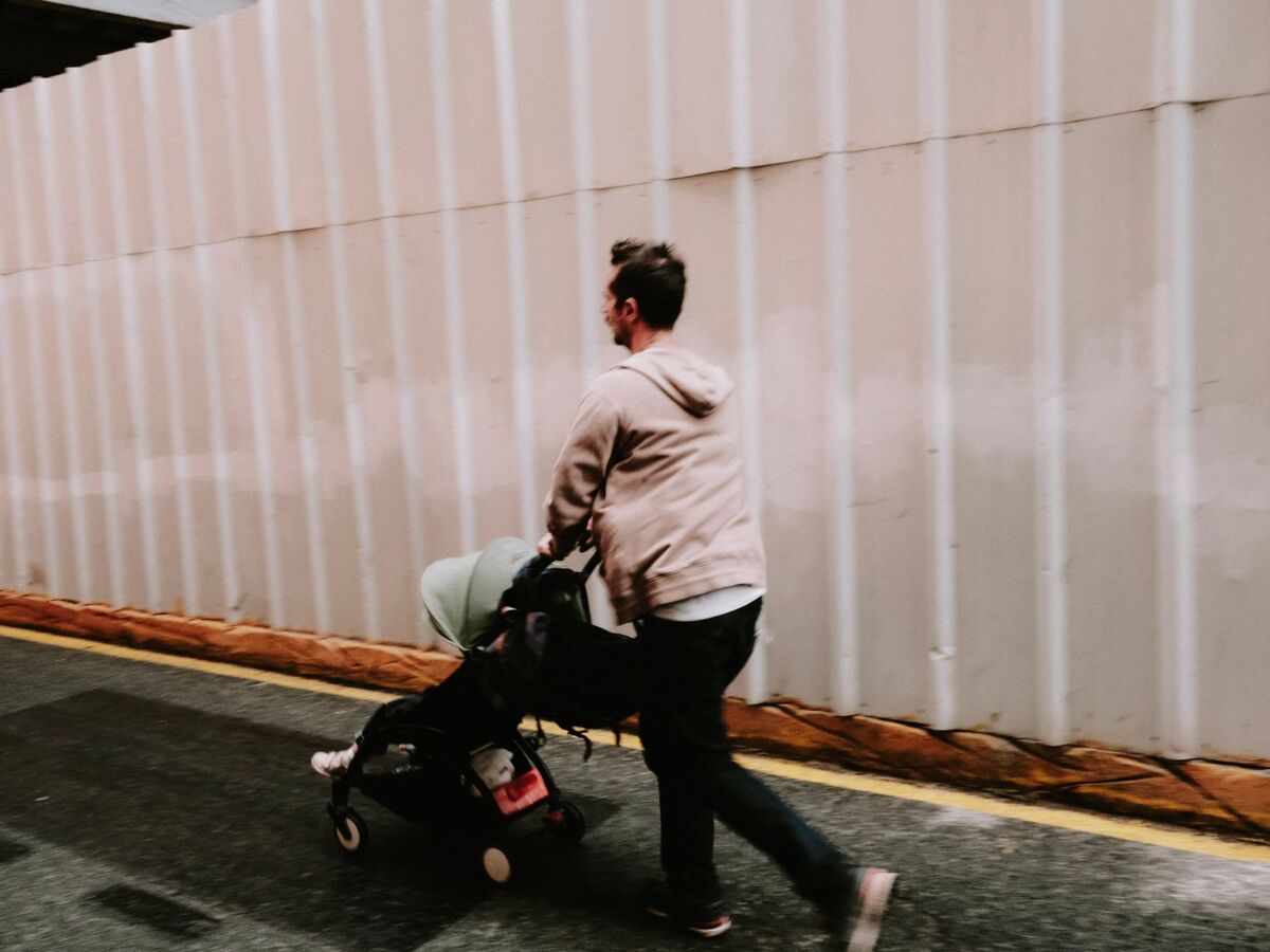 A man pushing a stroller down a street alongside a corrugated wall, captured in an urban setting.