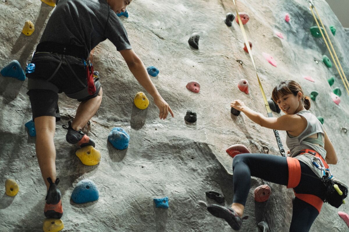 Back view of crop anonymous male trainer helping young Asian female athlete hanging on belay during climbing training in gym