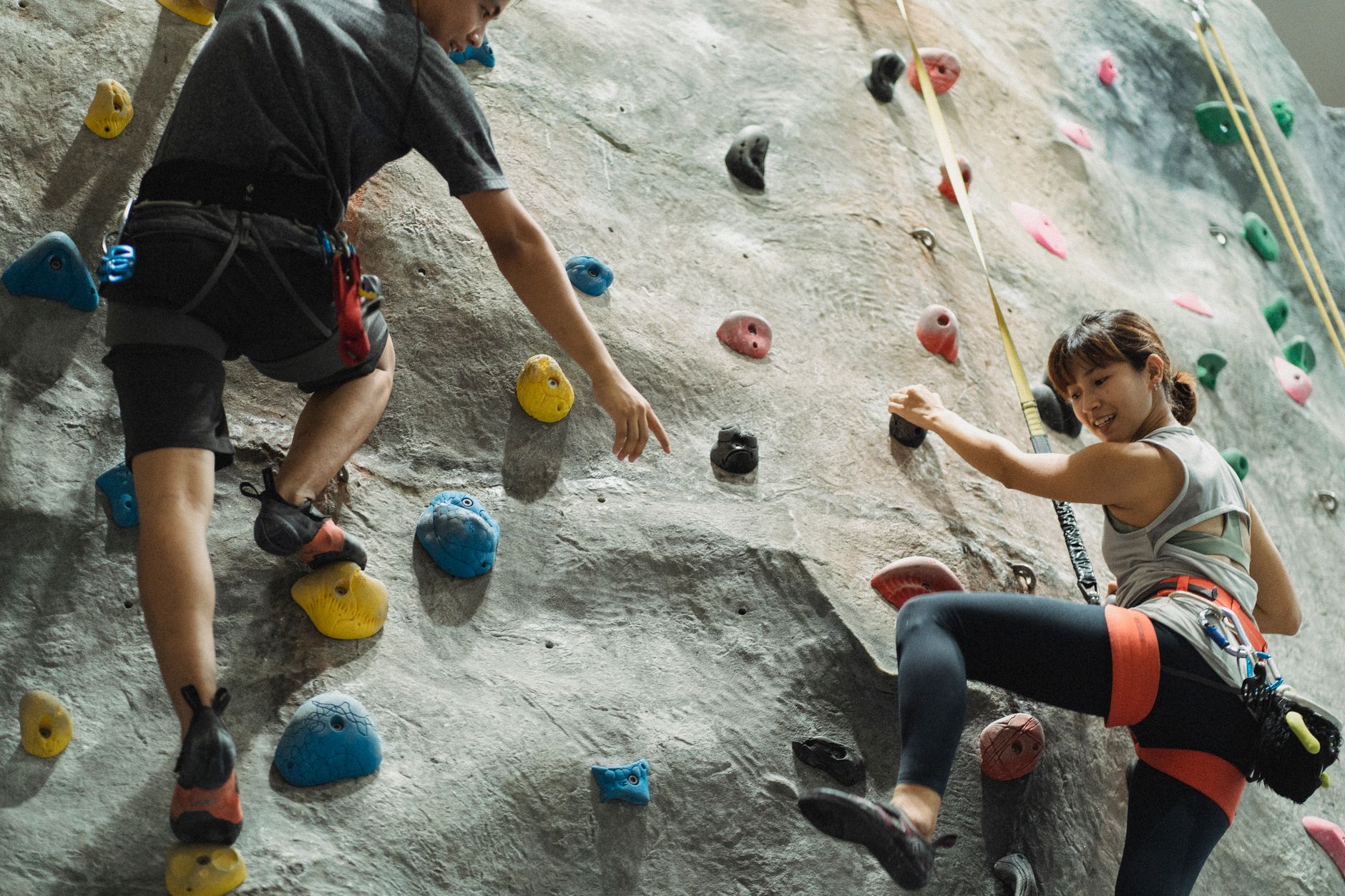 Back view of crop anonymous male trainer helping young Asian female athlete hanging on belay during climbing training in gym