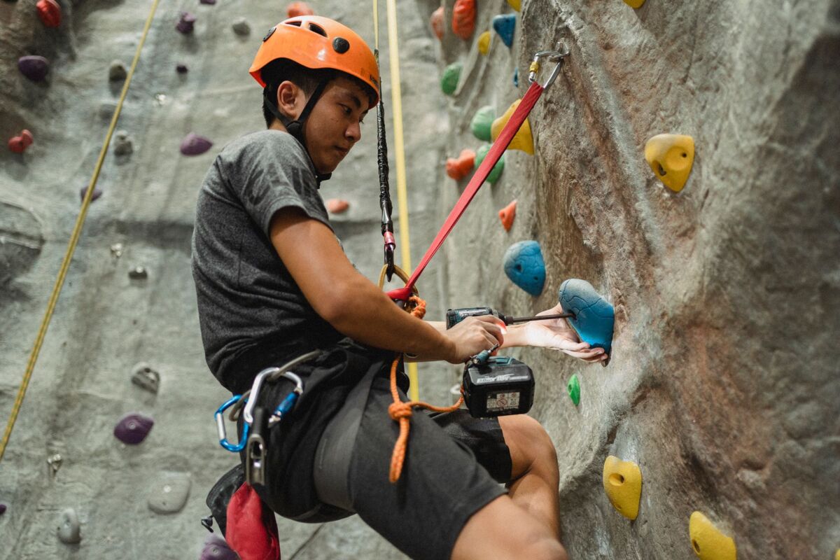 From below side view of attentive young Asian climber in protective helmet using electric screwdriver on hold of artificial rock