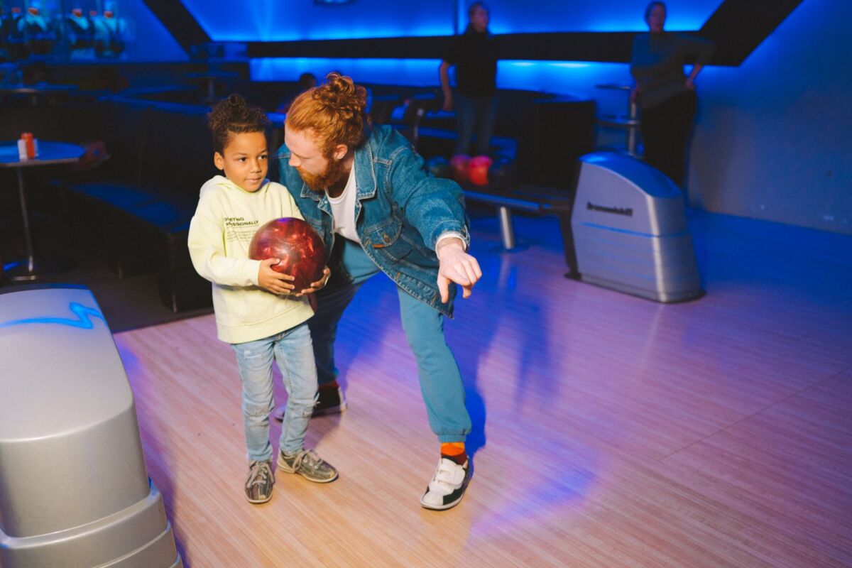 A bonding moment as a father teaches his young son to bowl at an indoor alley under blue lights.