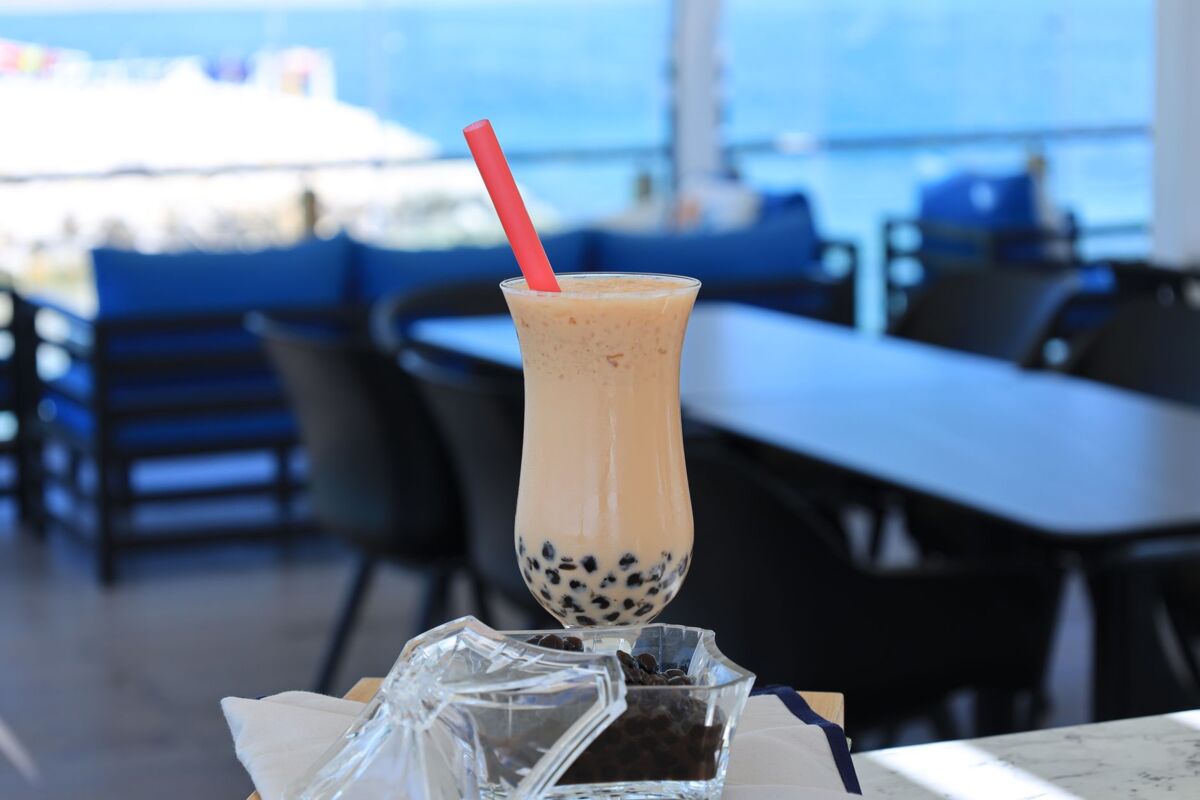 Chilled boba tea with tapioca balls and straw, set against a scenic seaside view in a modern cafe.