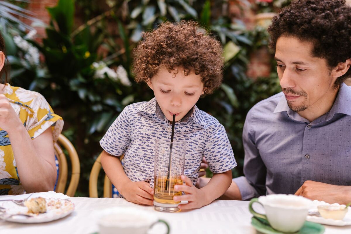 A child sipping a drink through a straw with father at an outdoor cafe table.