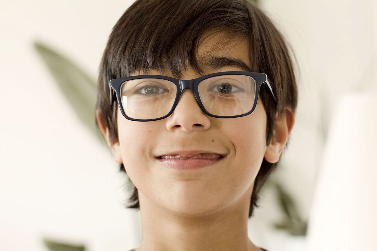 A close-up of a smiling young boy wearing eyeglasses indoors, captured in a natural light setting.