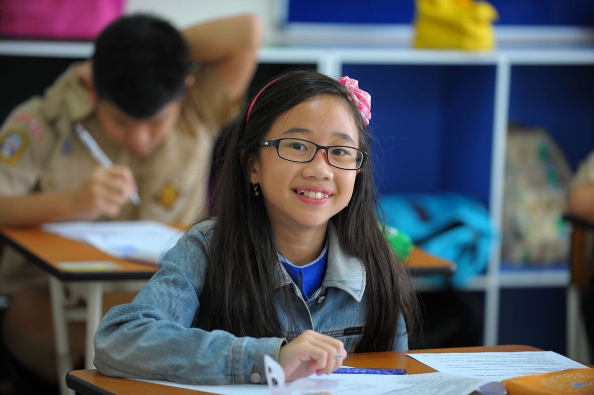 A young girl with glasses smiles while studying in a classroom setting.