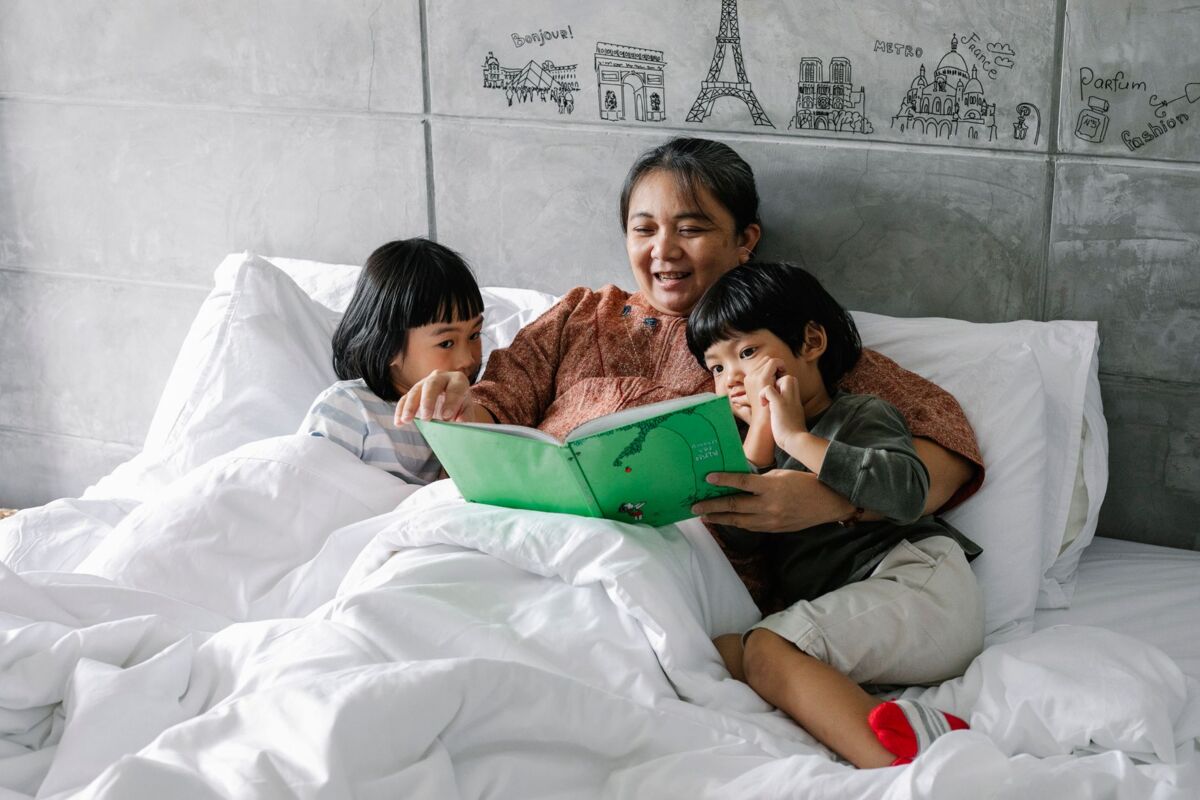 Positive aged ethnic grandmother smiling and reading book while lying on comfortable bed with cute attentive little grandchildren listening to story with intere