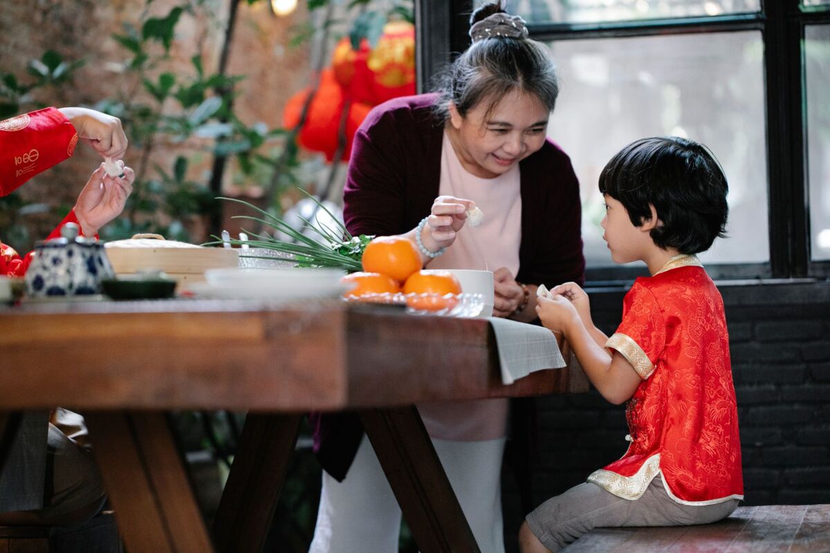 Positive elderly Asian female in casual clothes teaching adorable little grandson to fold traditional jiaozi dumplings during dinner preparation in kitchen