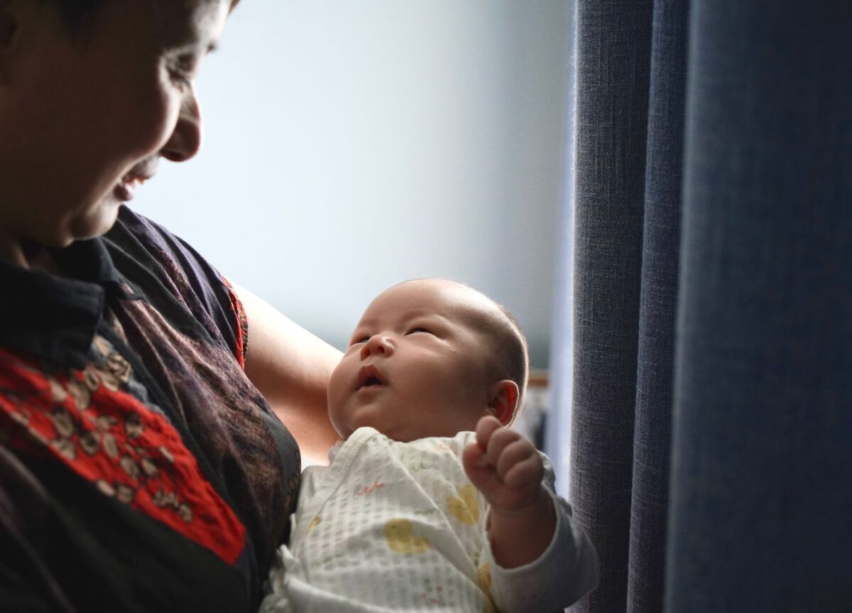 A touching moment between a grandmother and her newborn grandchild indoors near blue curtains.