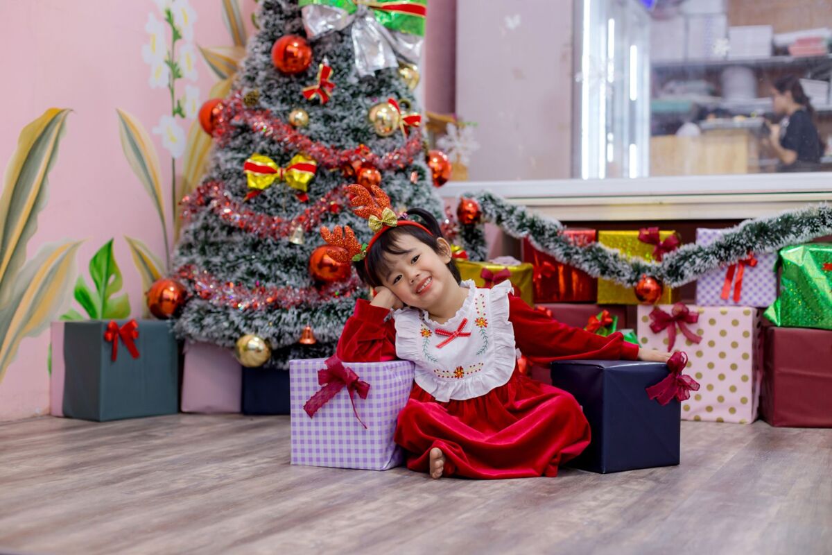 Happy child in festive attire sitting by a decorated Christmas tree in Hoi An, Vietnam.