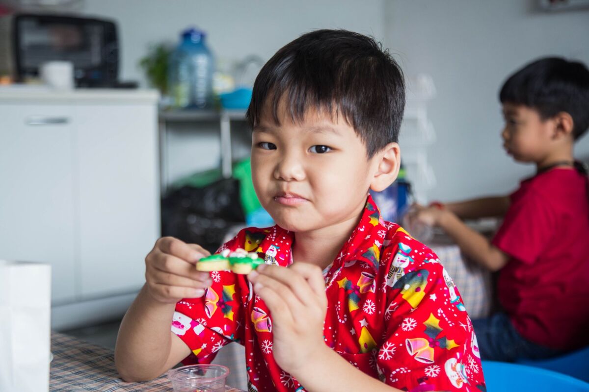 A young boy in a festive red shirt enjoys a colorful Christmas cookie indoors.