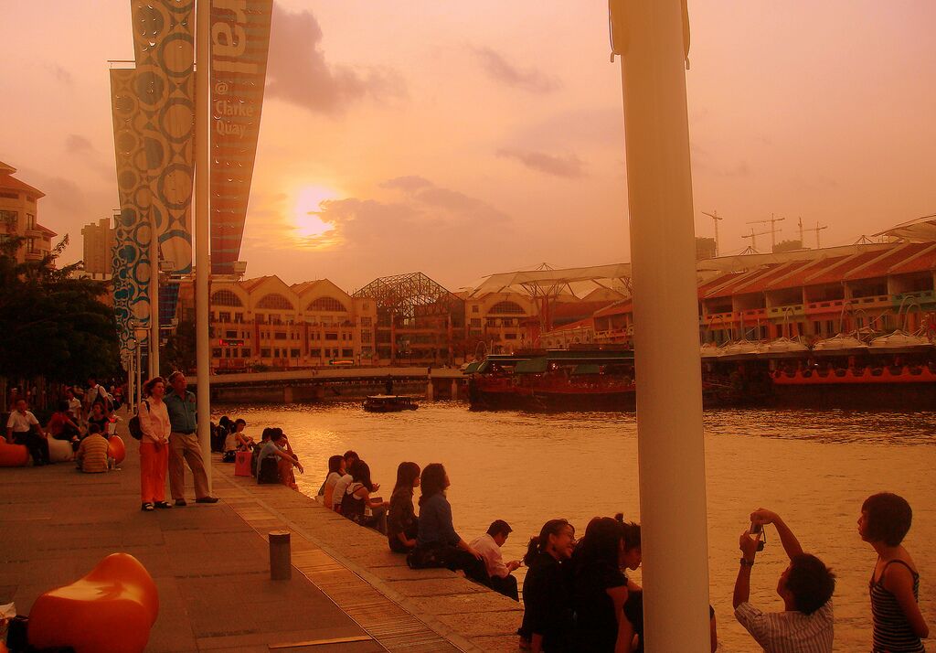 People sitting and walking along the Singapore River promenade opposite Clarke Quay at golden-hour sunset