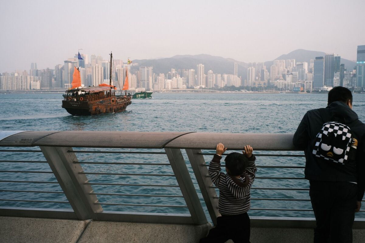 A father and child watch a traditional sailing ship in Hong Kong harbor with skyscrapers in the background.