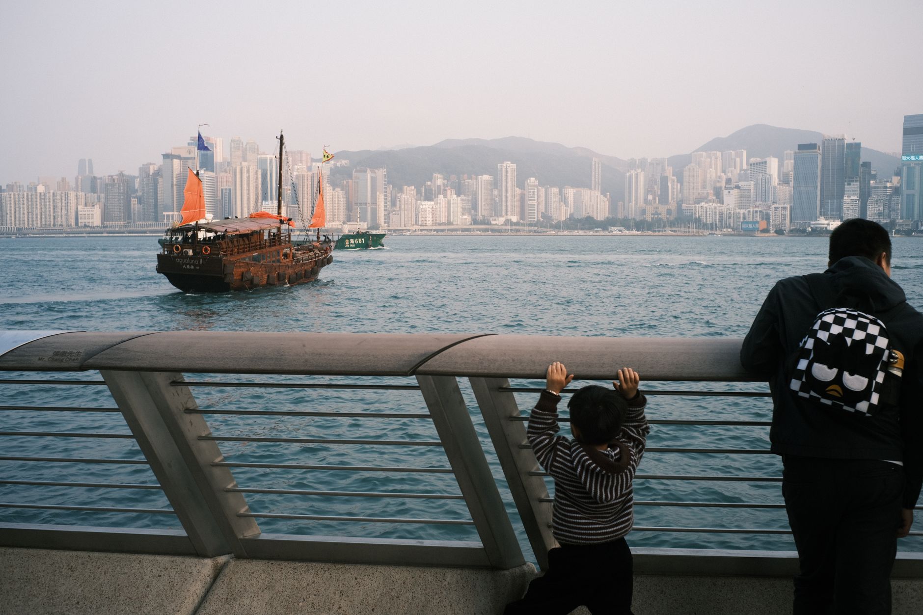 A father and child watch a traditional sailing ship in Hong Kong harbor with skyscrapers in the background.