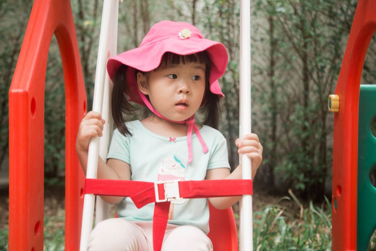 Cute Asian girl in a pink hat sitting on a swing at a playground.