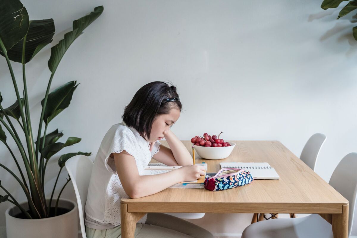 Asian child reading and writing at home, surrounded by plants and books, creating a learning environment.
