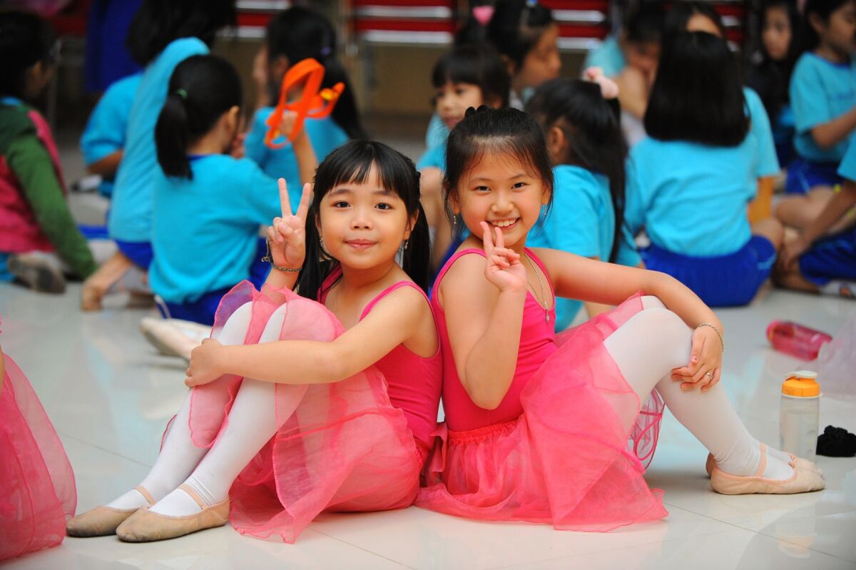 Two young girls in pink ballet tutus posing cheerfully indoors among peers.