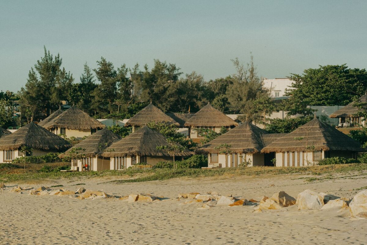 Scenic view of rustic beach huts with thatched roofs along a sandy shoreline, surrounded by lush greenery.