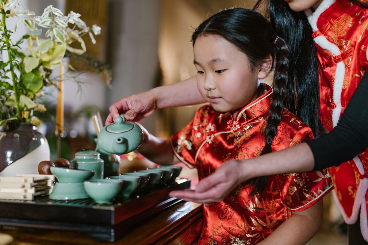 A young girl in traditional clothing learning a tea ceremony with her mother assisting, showcasing cultural heritage.
