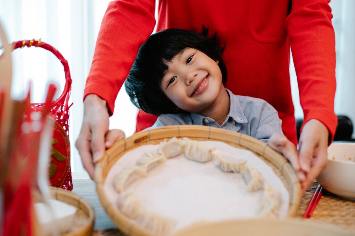 Cute positive ethnic boy showing bamboo bowl with raw homemade jiaozi while sitting near crop mother
