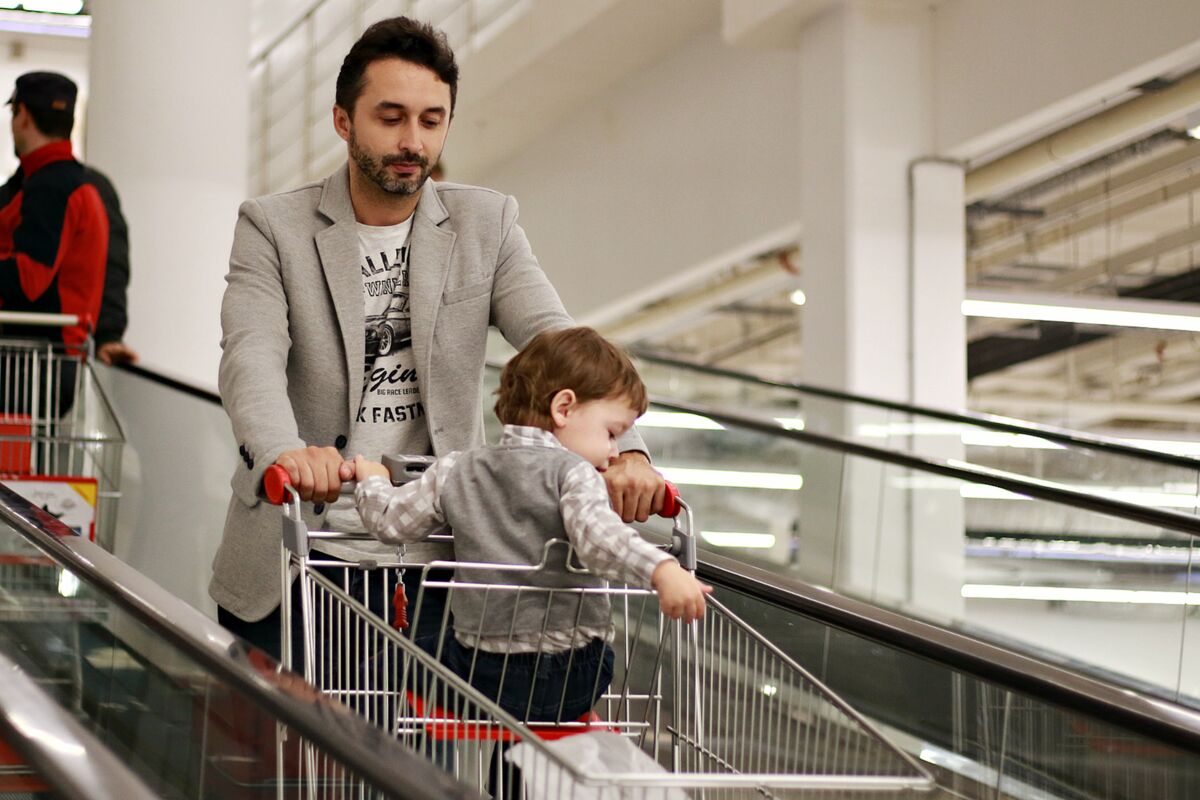 A man with a child in a shopping cart using an indoor escalator at a mall.