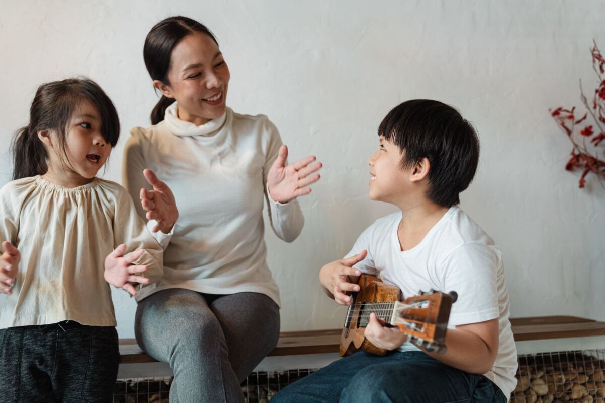 Happy ethnic mother and daughter clapping hands and singing song while positive son playing Hawaiian musical instrument at home